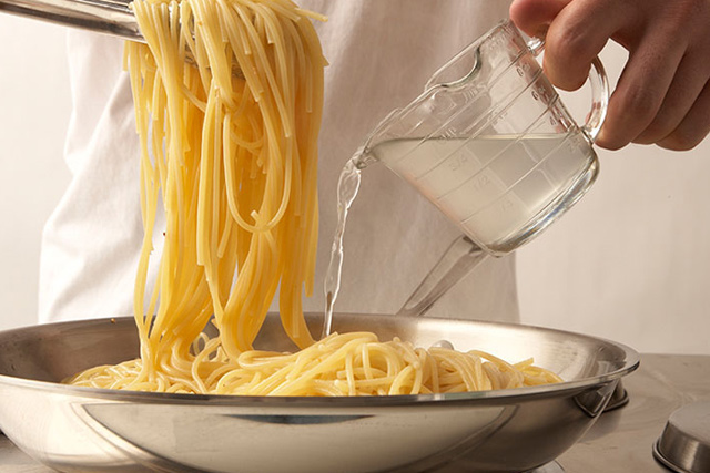 Eine Person gießt Wasser über Spaghetti in einem Topf auf einem Herd.